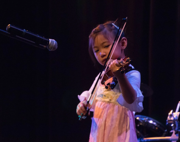 Young person playing a violin on stage, facing forward. Stage has a microphone and dark backdrop.