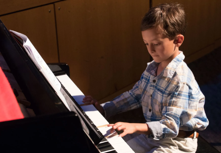 Young boy playing piano, lit by sunlight. He wears a plaid shirt, focused on the keys.