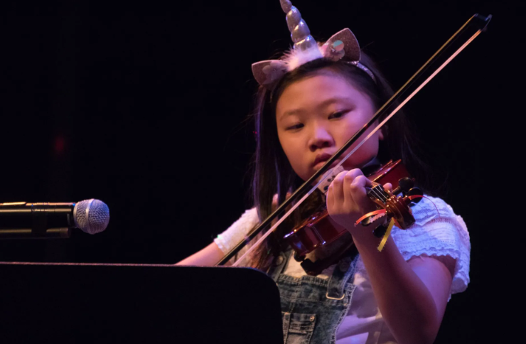 Young girl with unicorn headband playing violin on stage near microphone.