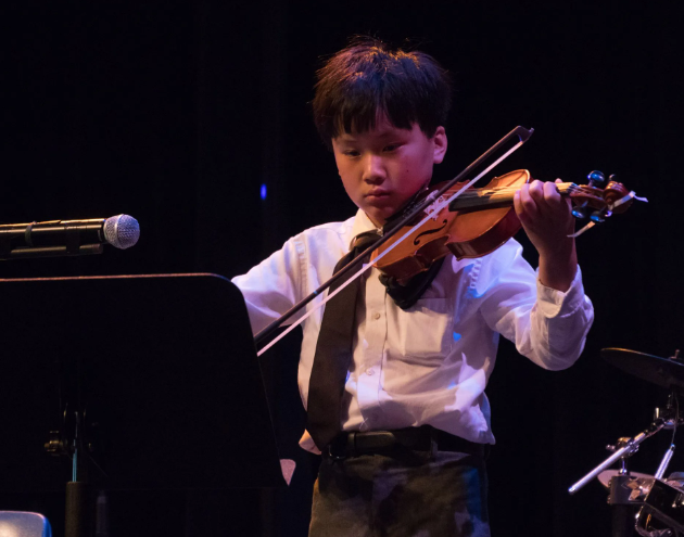 Young person playing a violin on stage, wearing a white shirt and black tie, with a music stand nearby.