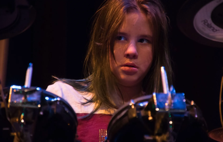 Young person playing electronic drums, focused expression, dark setting.