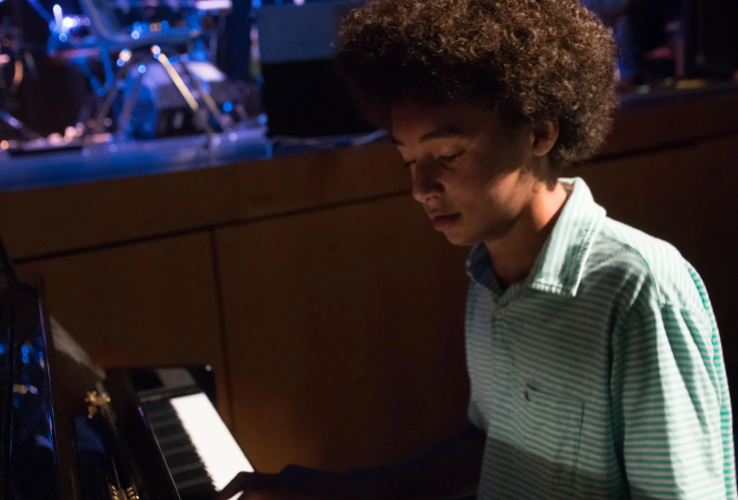 Teen playing a piano with a band onstage, wearing a light blue striped shirt.