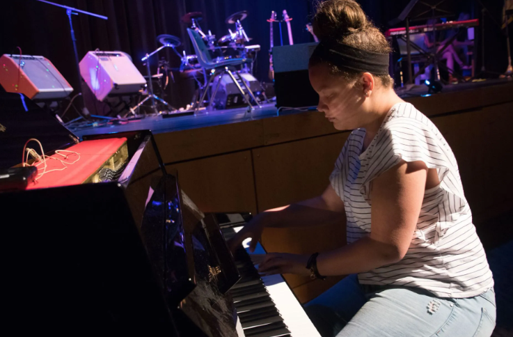 Woman playing a black piano on stage. Band instruments are in the background, blue stage lighting.