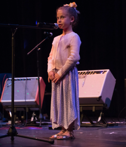 Young person on a stage, speaking into a microphone, with a keyboard and amps in the background.