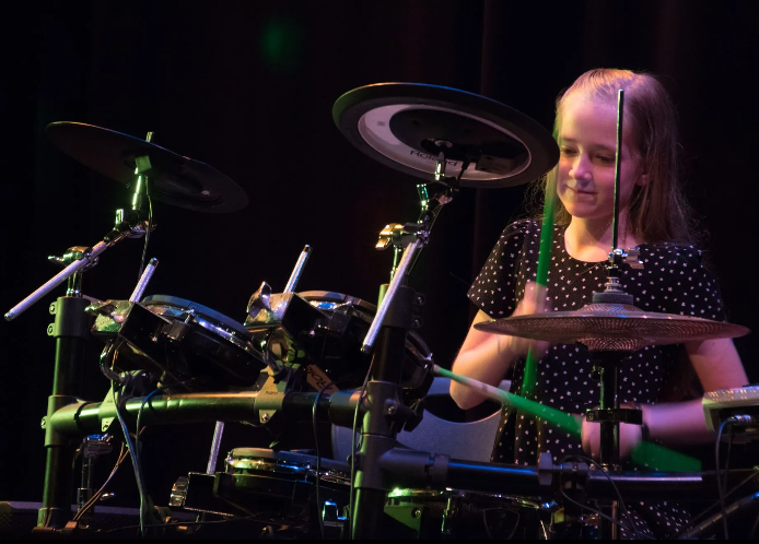 Young person playing electronic drums on stage, smiling with green drumsticks.