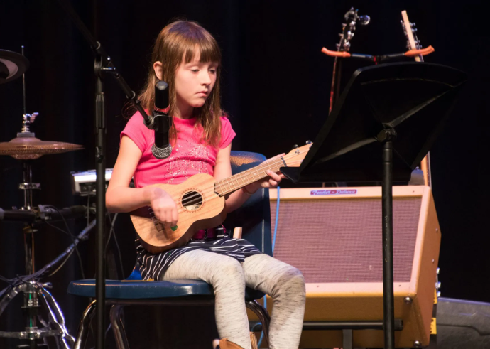 Girl playing a ukulele on stage, reading music from a stand. She wears a pink shirt.