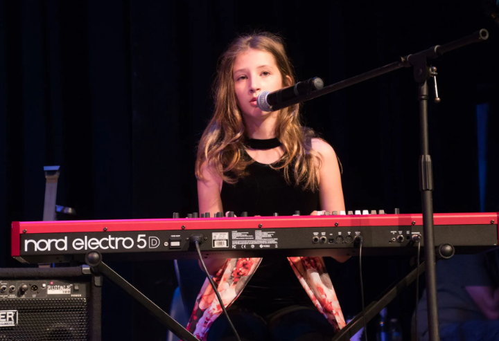 Young person playing a red Nord keyboard and singing into a microphone on stage, black background.