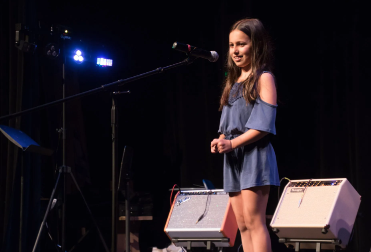 Girl on stage with a microphone, wearing a blue romper. Stage with equipment, dark background.