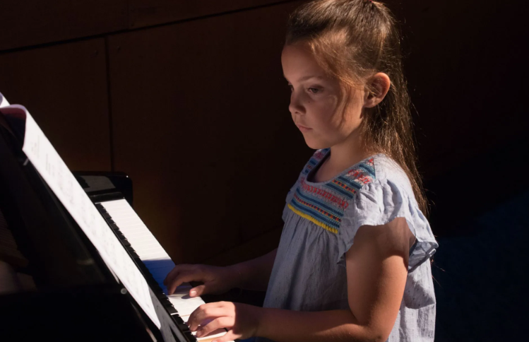 Girl playing piano, focused on sheet music, lit by sunlight.