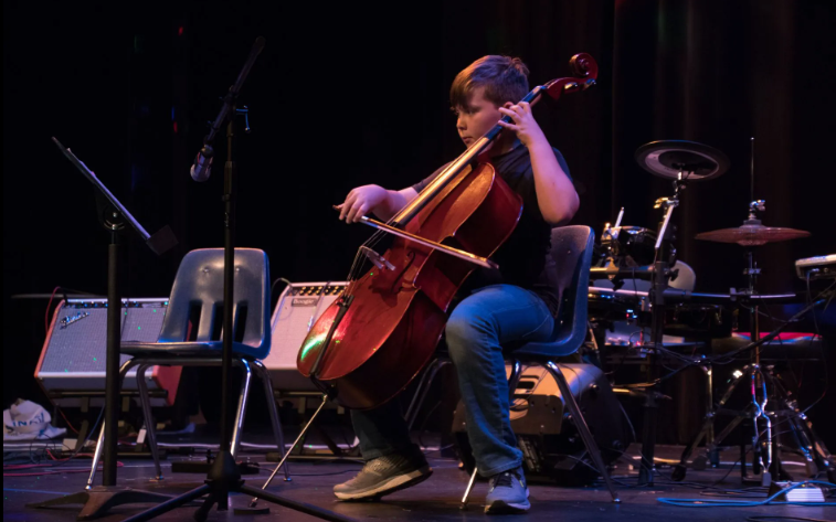 Person playing a cello on a stage, lit with stage lighting. They are seated, with a bow in hand.