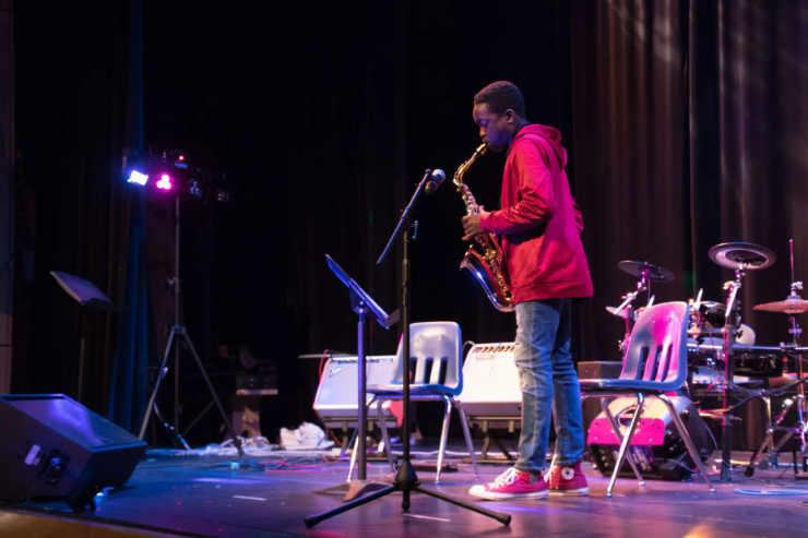 A person in a red jacket plays a saxophone on a stage, with drum set and sound equipment visible.
