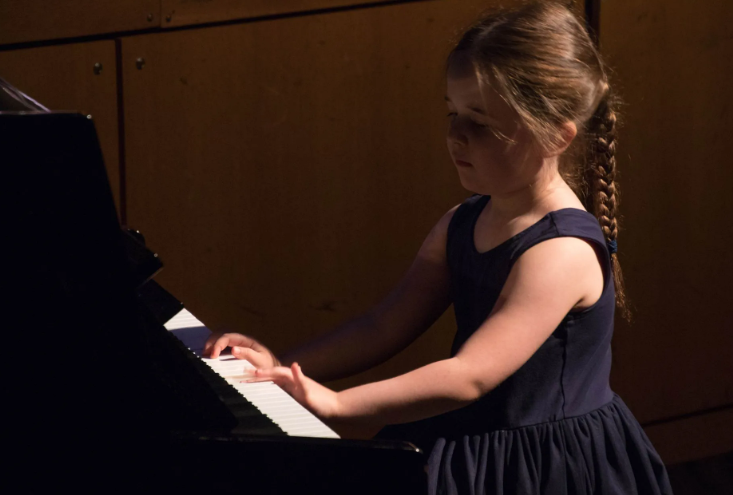 Girl in a navy dress playing a black piano, side view. She has a braid and is focused on the keys.
