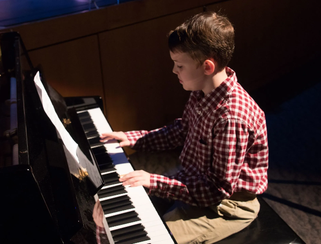Boy playing a piano, wearing a plaid shirt, hands on keys, indoors.