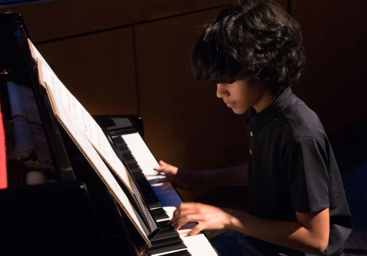 Boy playing a grand piano, reading sheet music. Dark room, focused expression.