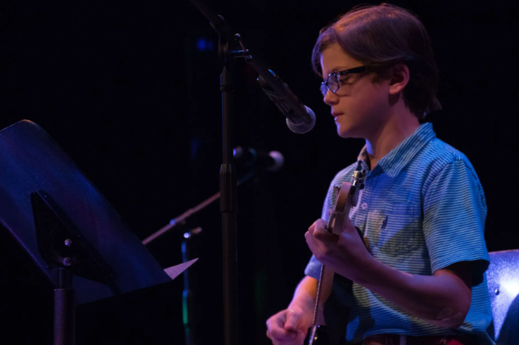 Boy playing electric guitar onstage with microphone and music stand.