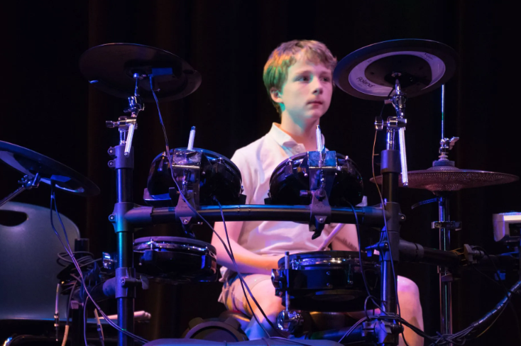 Boy playing an electronic drum set on stage. Dark setting, blue lighting.