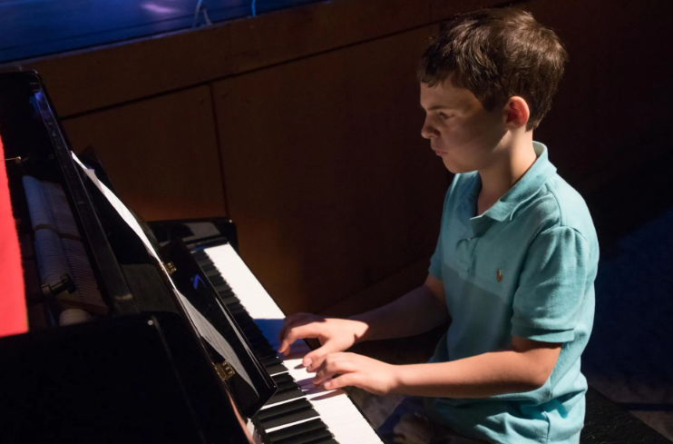 Boy playing a piano in a dimly lit room, wearing a blue polo shirt.