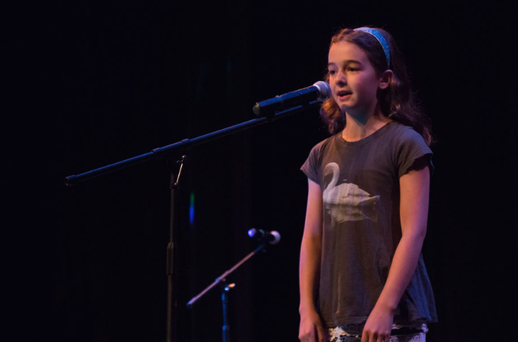 Girl singing at a microphone on a dark stage, wearing a gray shirt with a swan graphic.