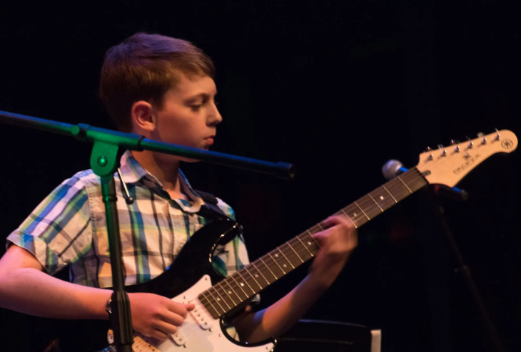 Boy playing an electric guitar on stage, lit by spotlights.