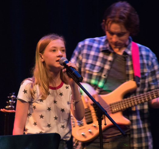 Girl singing into a microphone onstage; a bass guitarist is behind her.