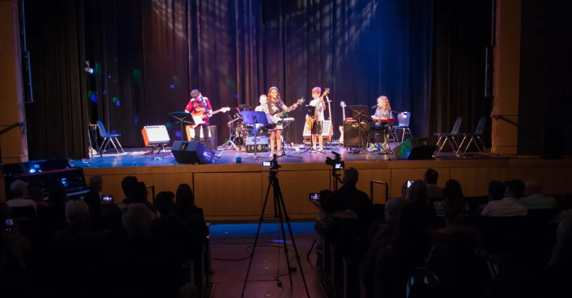 A band performs on a stage with a seated audience in front. Stage lights shine on performers with instruments.