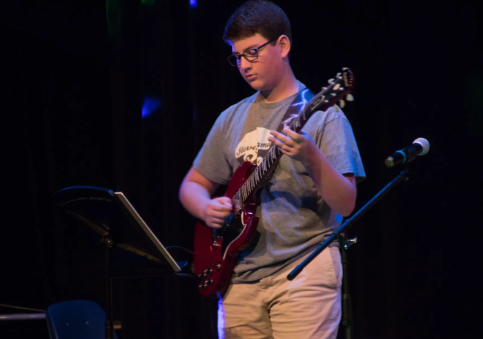 Person playing a red electric guitar on a stage with a microphone. He wears glasses and a gray t-shirt.