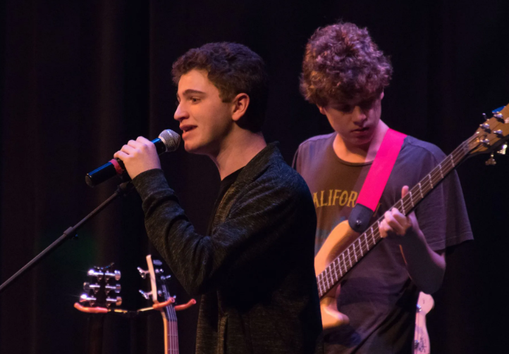 A singer performs on stage with a microphone; a bassist plays guitar behind him. Dark setting.