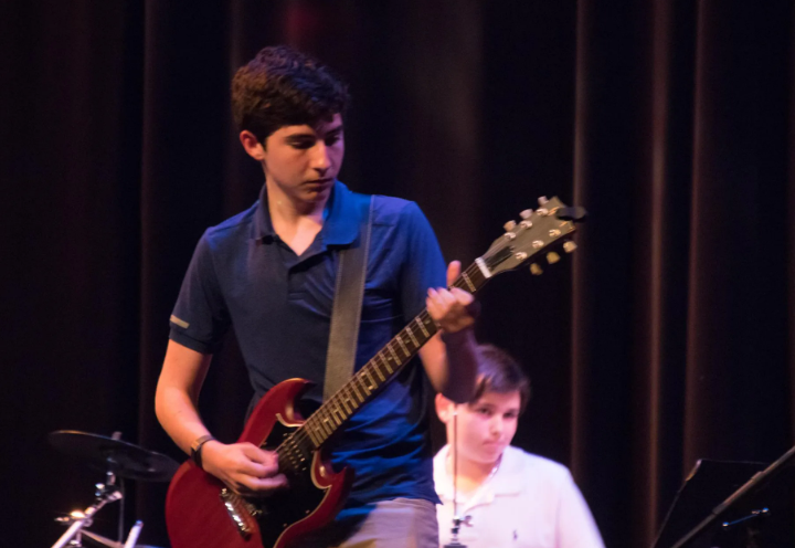 Teen plays red electric guitar on stage; another person in background.
