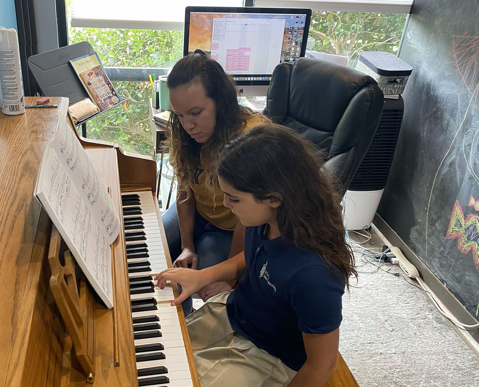 Woman helps a child play piano, both looking at sheet music. Indoors, near a computer and air cooler.