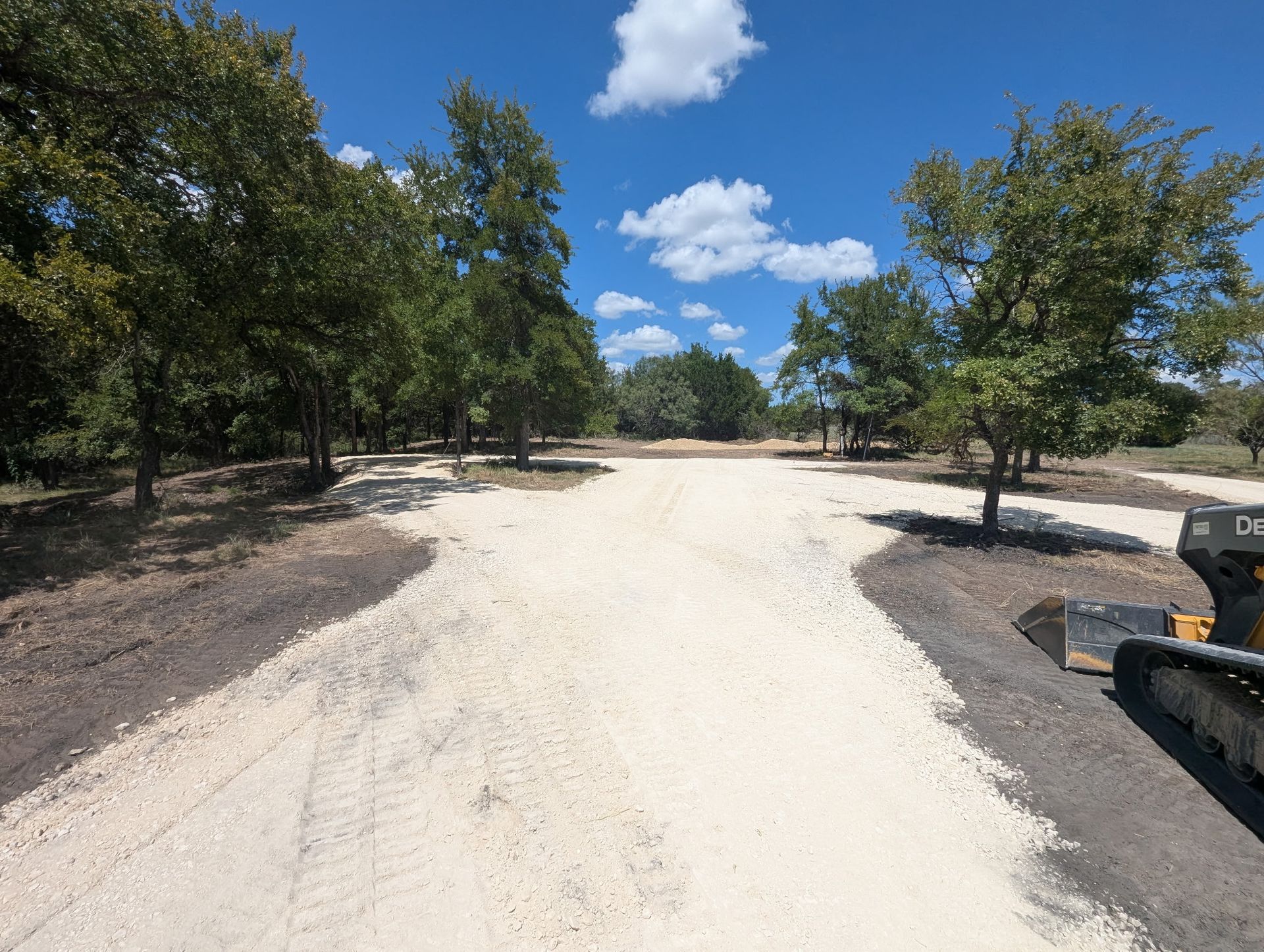 Gravel driveway surrounded by trees under a blue sky with fluffy clouds.