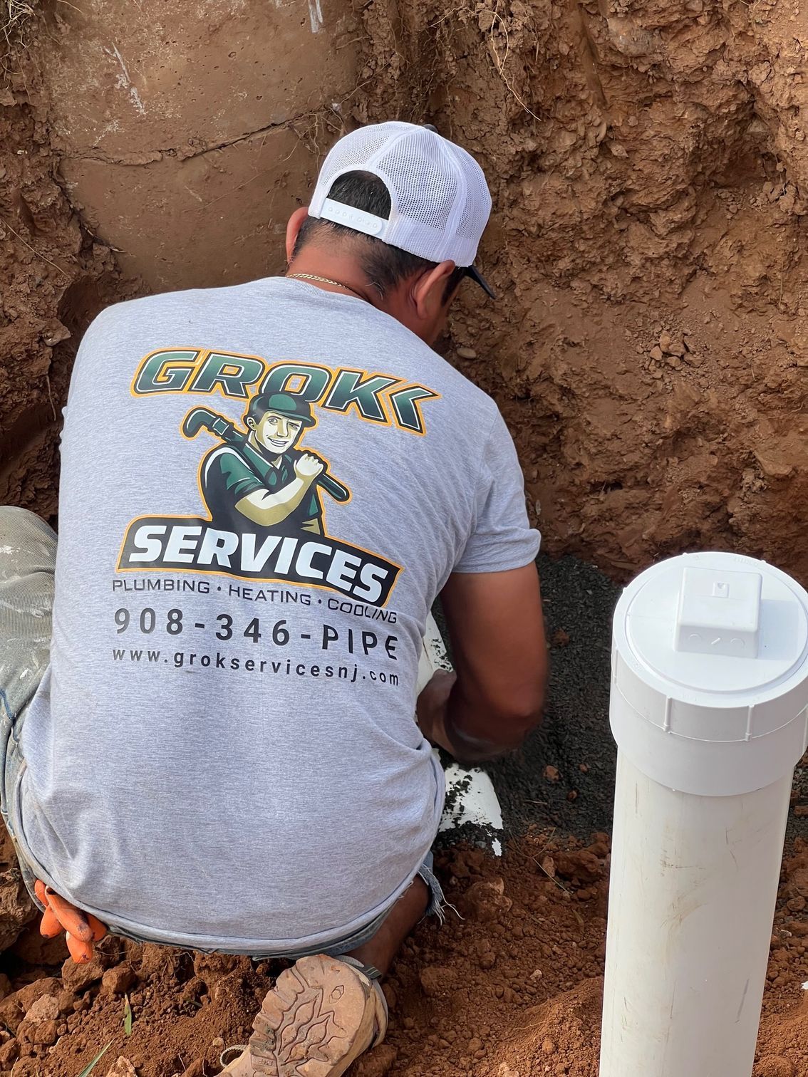 plumber in grey shirt working on a white pipe in a dirt trench