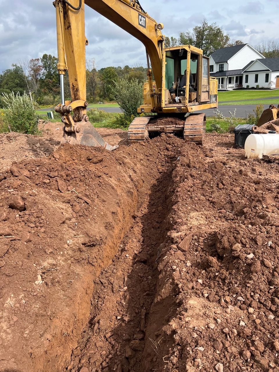 yellow excavator digging a trench in brown soil, next to a field with a white house in the background