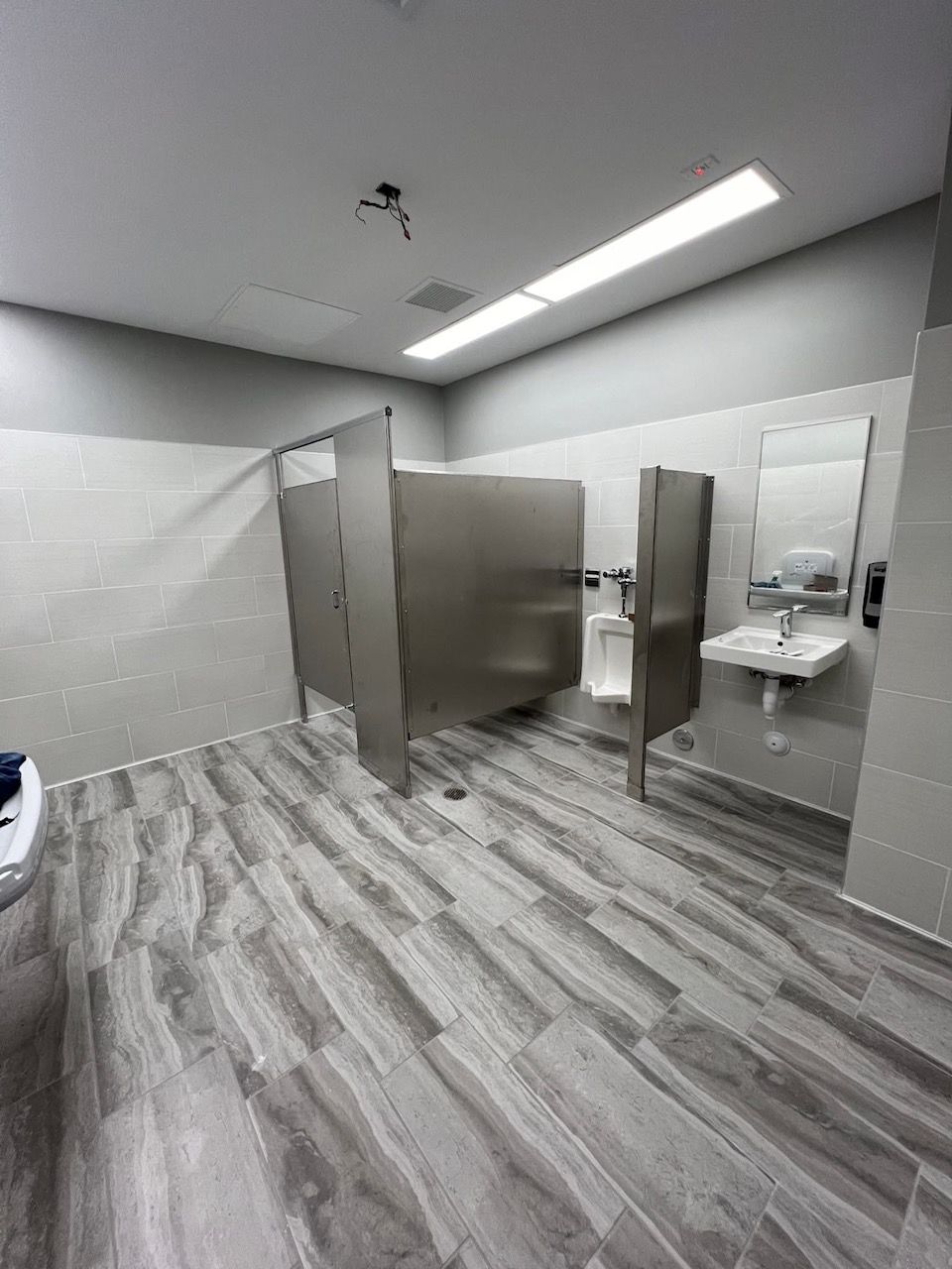 public restroom with stainless steel stalls, urinal, sink, and gray tile flooring