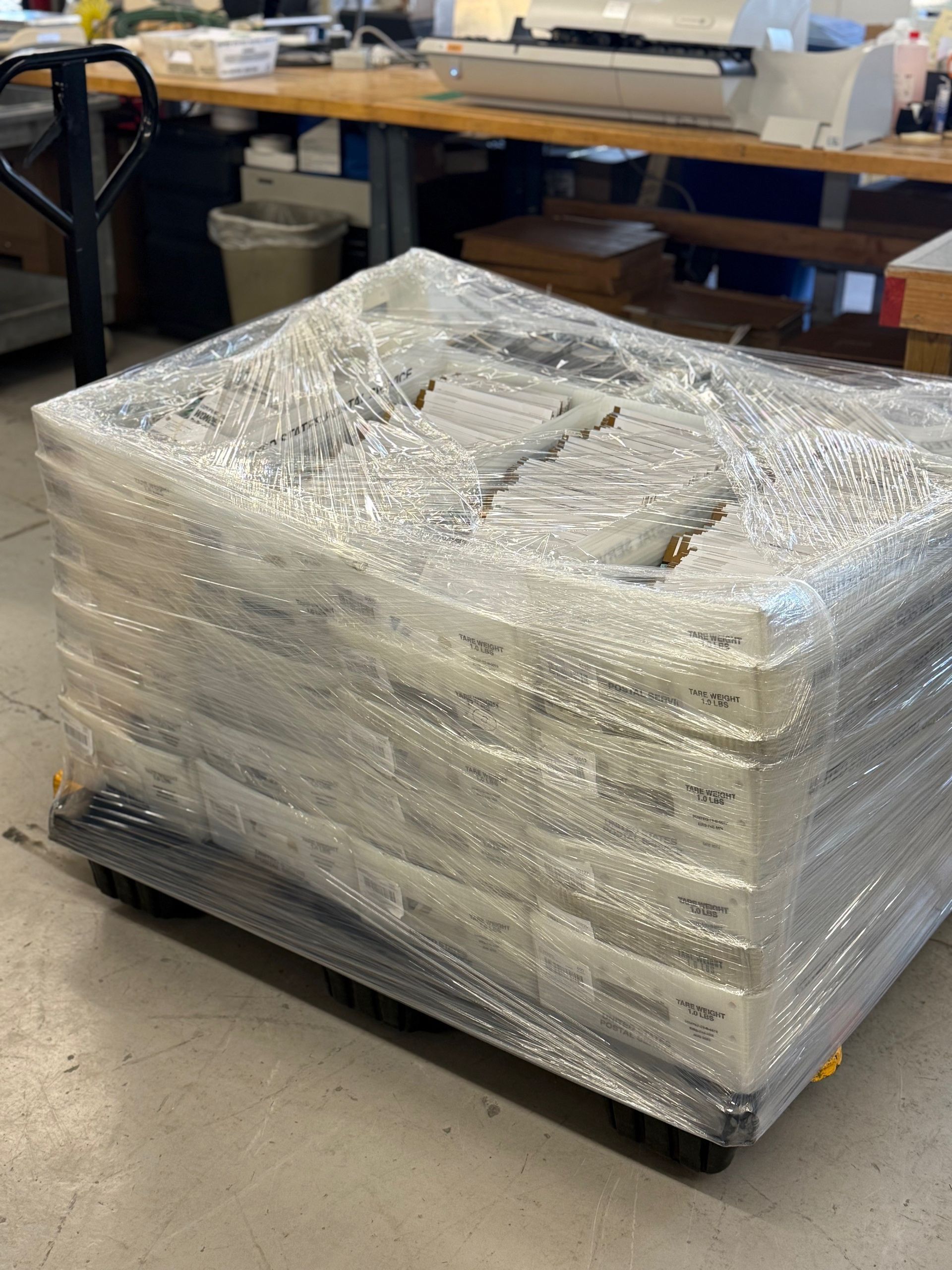 Pallet of white boxes wrapped in plastic, on a cart in a workshop setting.