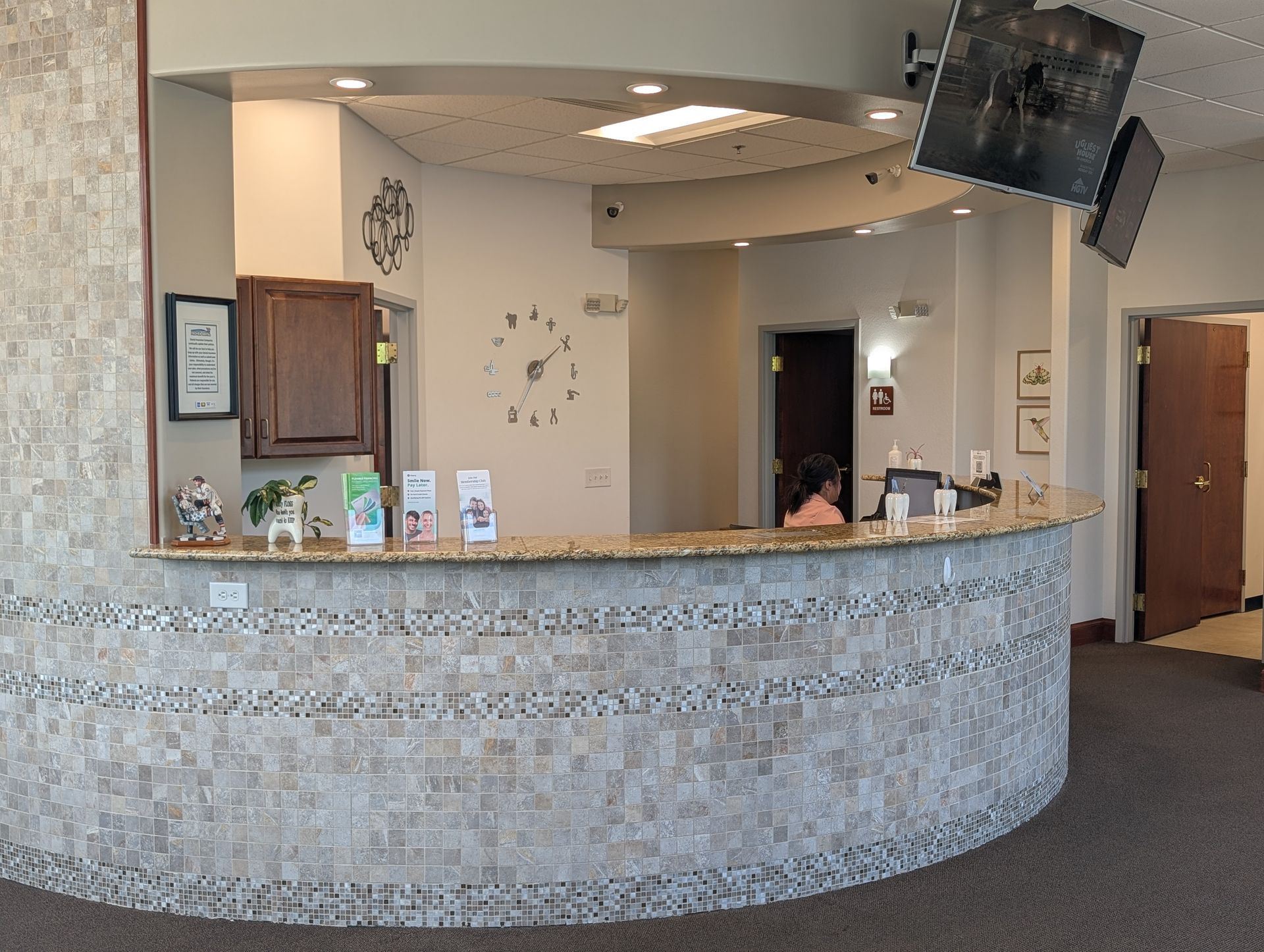 A woman is sitting at a counter in a dental office with a clock on the wall.