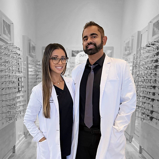 A man and a woman in white medical coats standing in an optical store with glasses on shelves on either side.