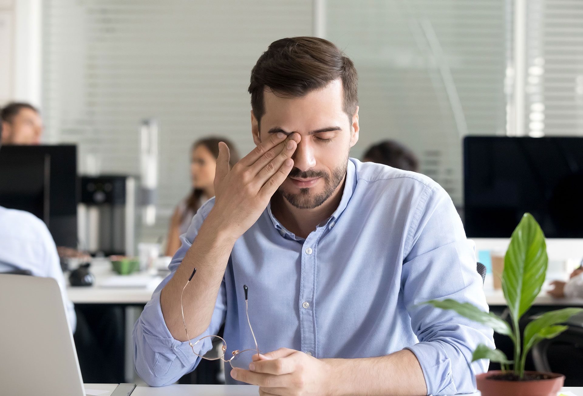 A man is rubbing his eyes while sitting at a desk in an office.