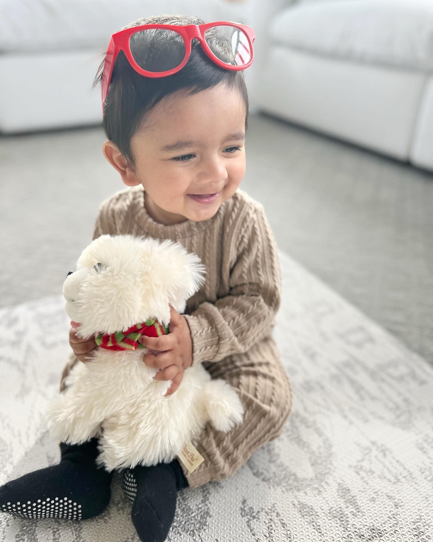 A baby wearing sunglasses is sitting on the floor holding a stuffed animal.