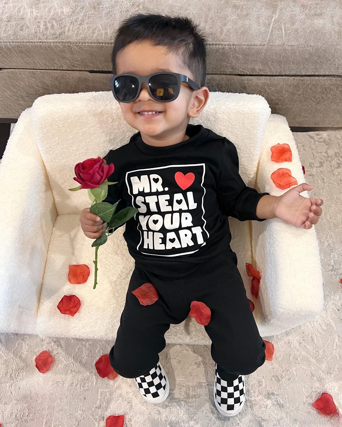 A little boy wearing sunglasses is sitting in a chair holding a rose.