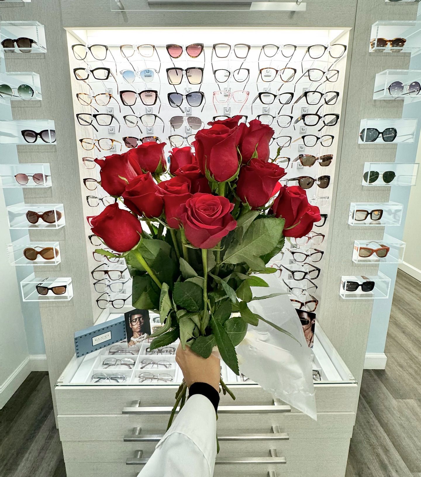A person is holding a bouquet of red roses in front of a display of glasses.