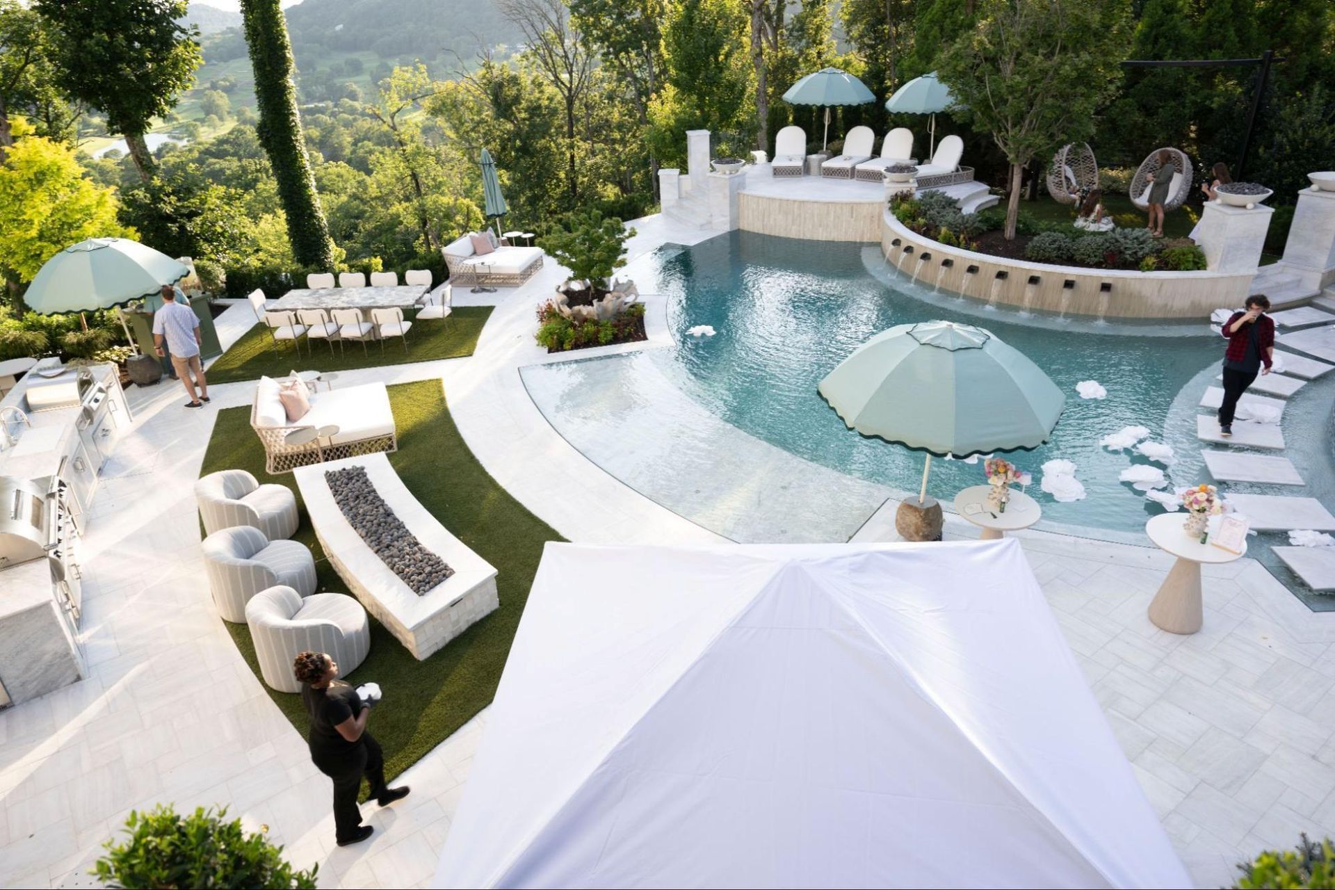 Luxury outdoor event: Pool, lounge areas, white umbrellas, lush landscaping, and people enjoying the sunny day.