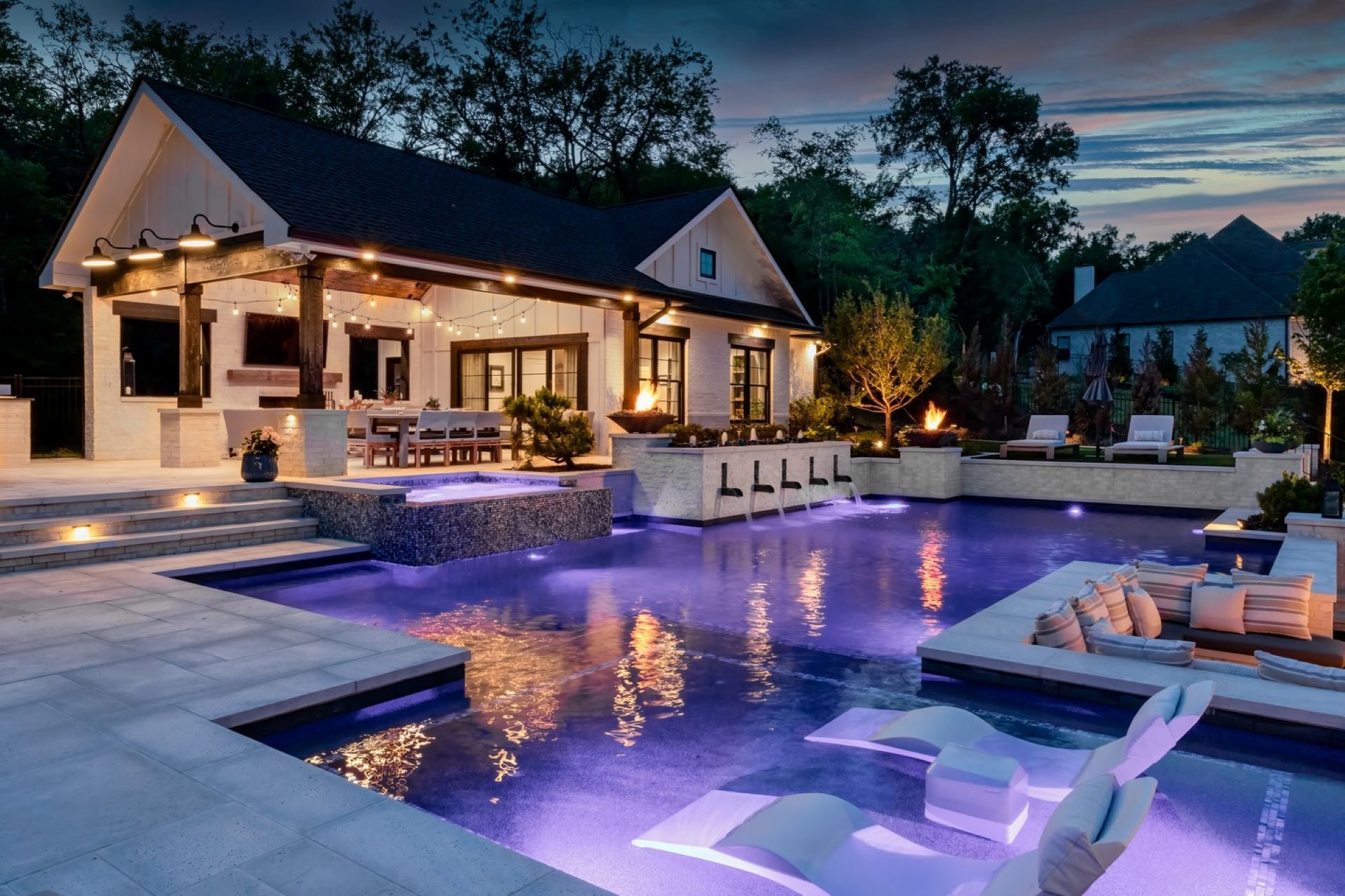 Night scene of an illuminated pool area with a covered patio, lounge chairs, and a small building.