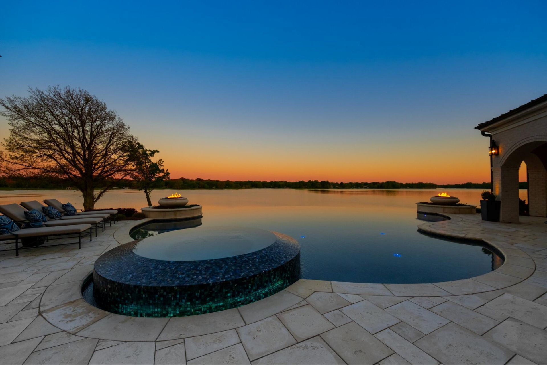 Infinity pool overlooking a lake at sunset. Features include a hot tub, lounge chairs, and a stone patio.