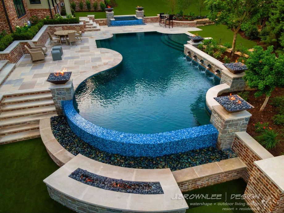 Luxury backyard pool with blue-tiled edges, waterfalls, fire features, and stone patios, surrounded by grass and landscaping.
