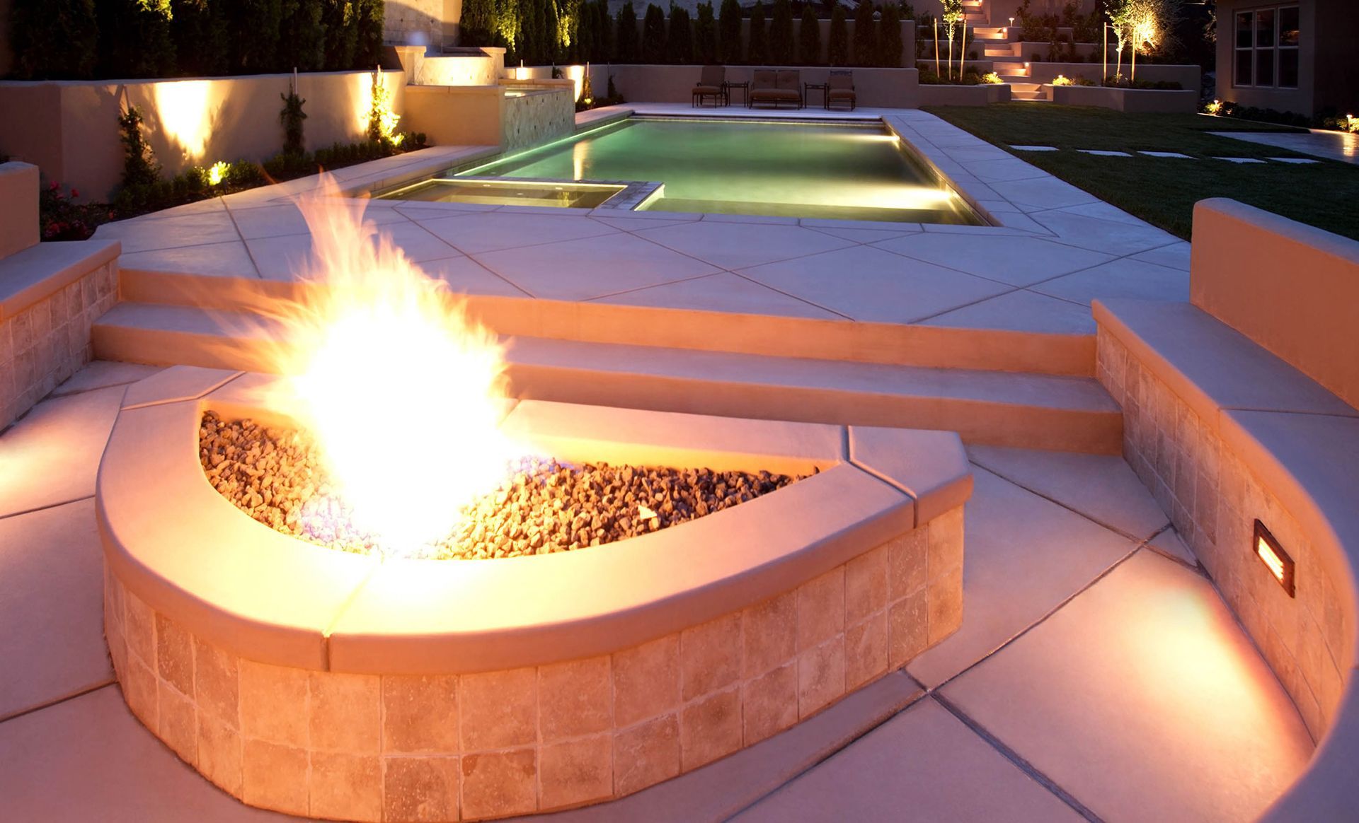 Fire pit with flames in a modern outdoor setting with a pool and built-in benches, lit at night.