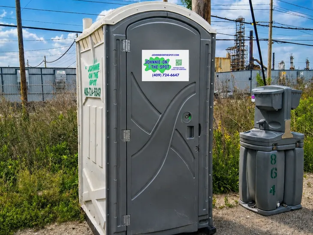 Gray portable toilet and handwashing station beside a fence at an industrial site.
