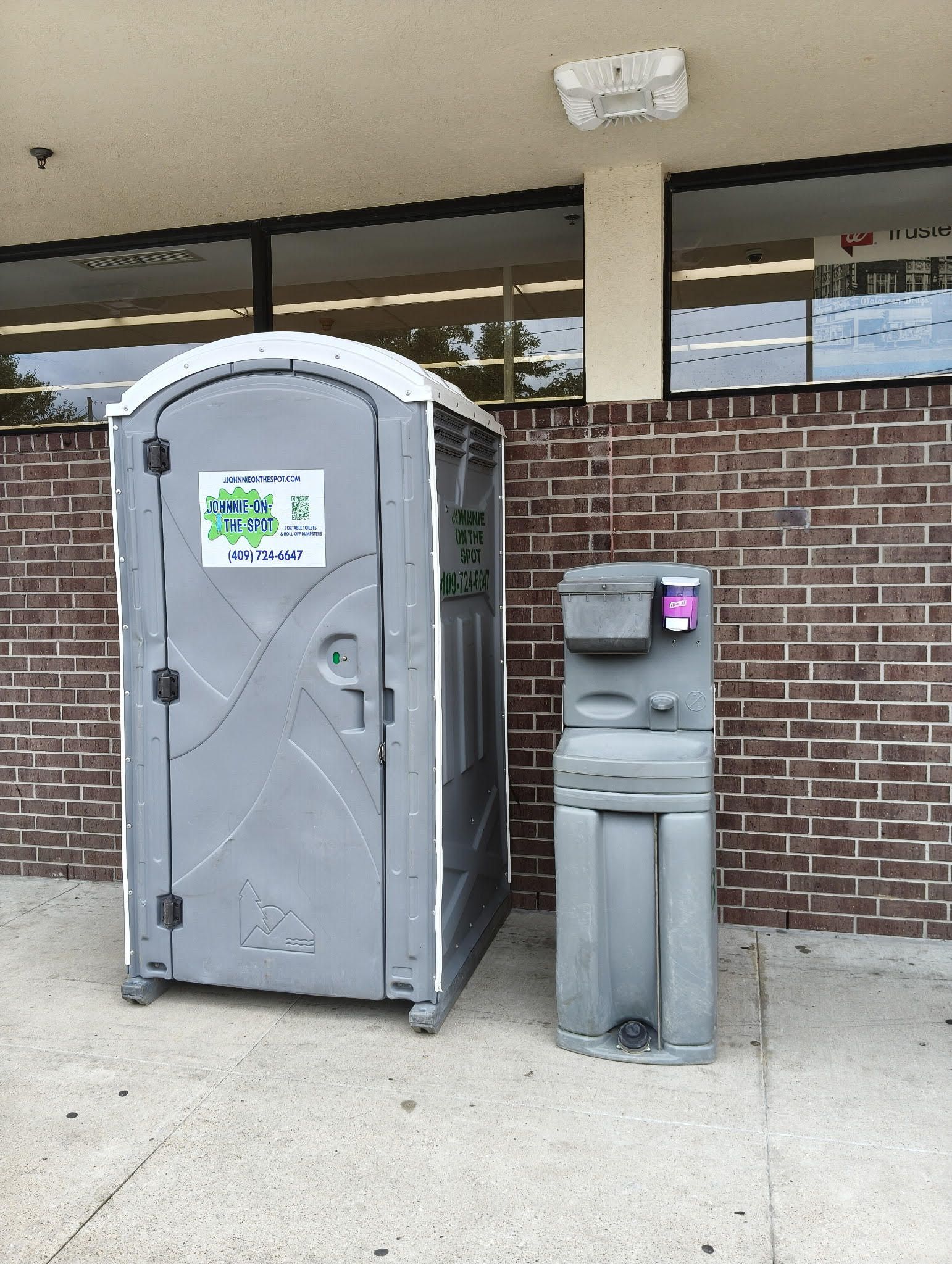 Portable toilet and handwashing station beside a brick wall outdoors.