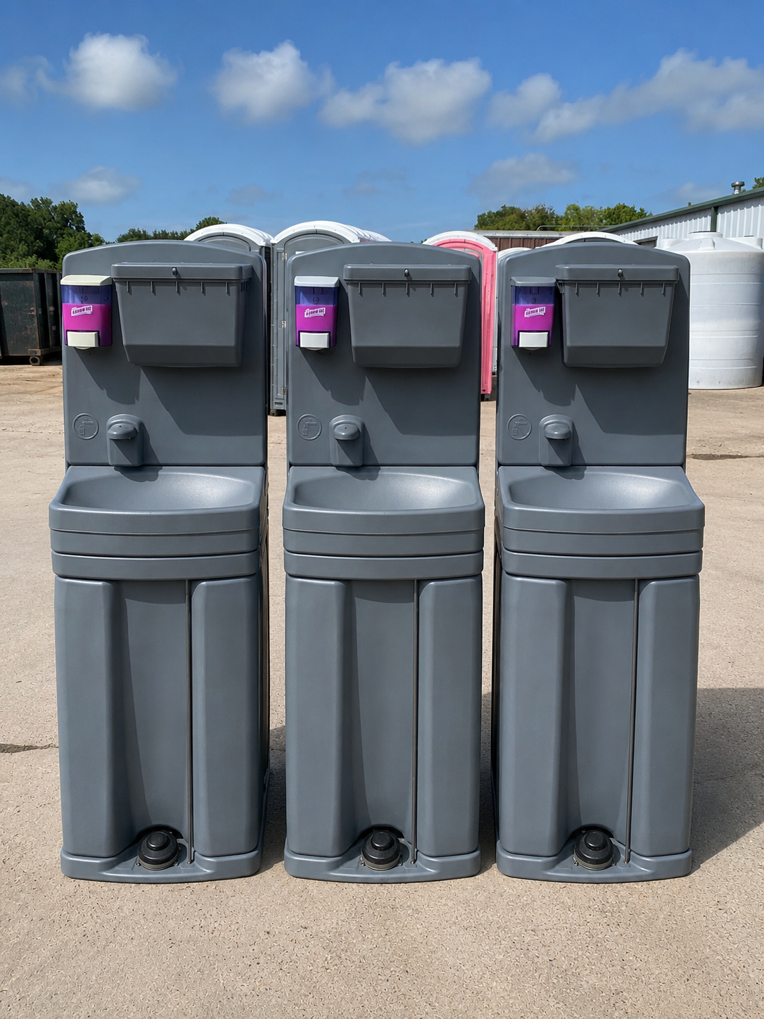 Three gray portable toilets lined up outdoors on a paved lot under a blue sky.