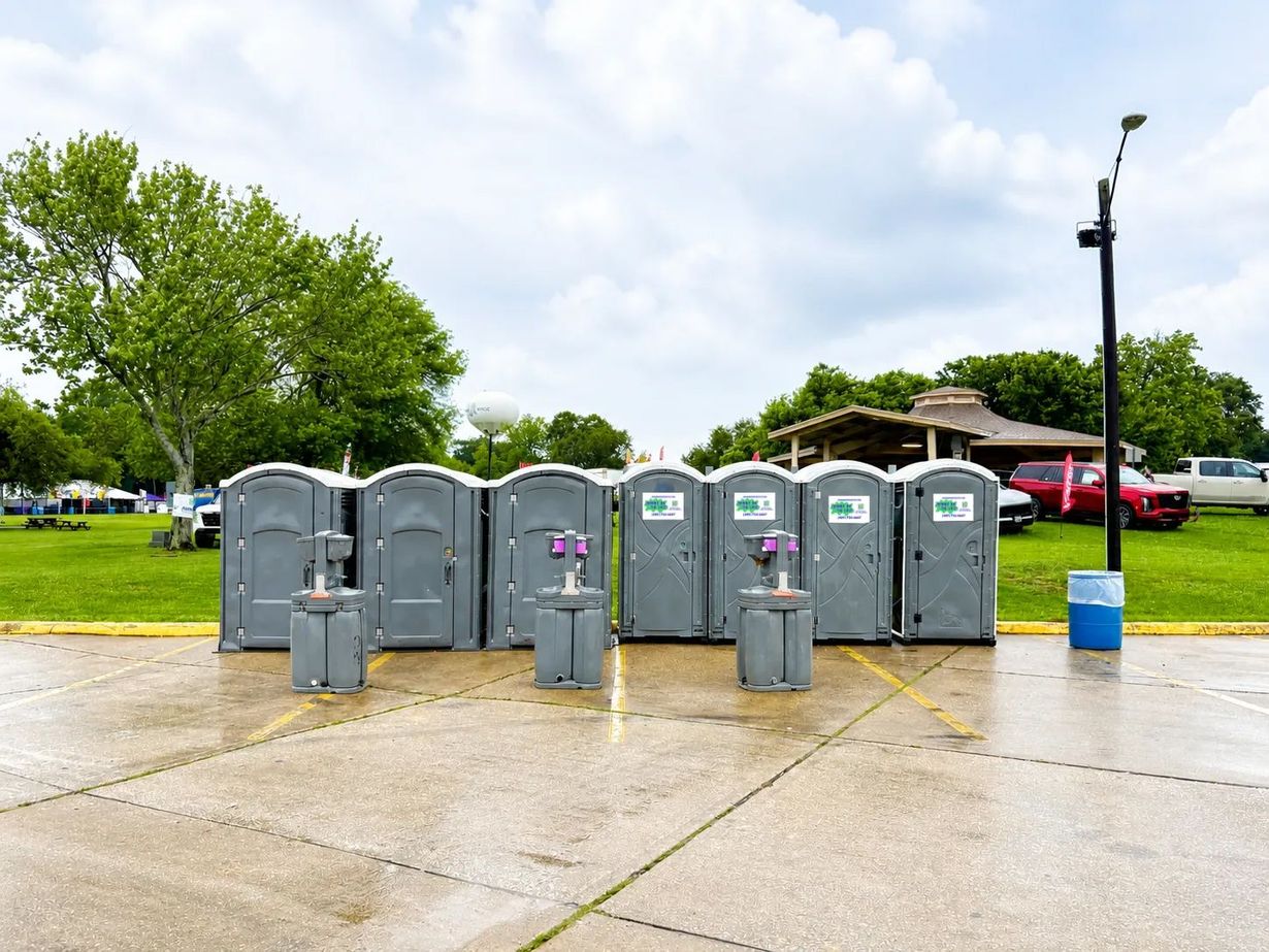 Row of gray portable toilets at a park, with a blue trash can and trees in the background.