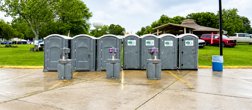Gray portable toilets lined up on a wet sidewalk beside a grassy park and parked cars, under cloudy skies